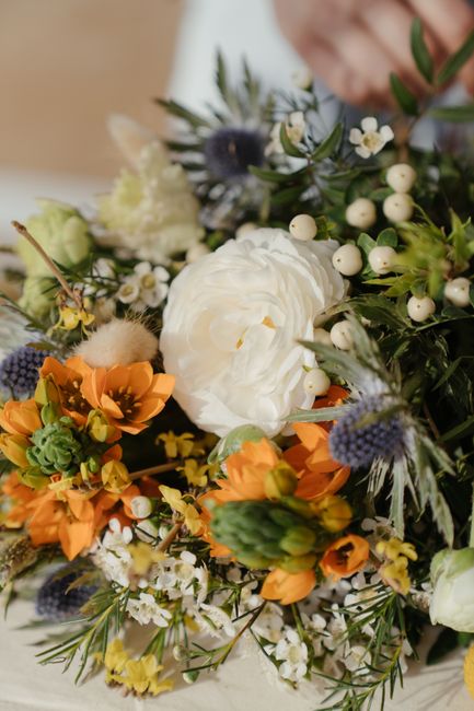 A vibrant floral arrangement featuring a large central white ranunculus with tightly layered petals, surrounded by bright orange flowers, small white blossoms, and sprigs of green foliage. Intersperse