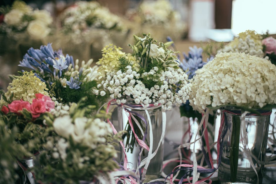 A collection of fresh floral arrangements featuring white hydrangeas, pink roses, blue lilies, and delicate white baby's breath, arranged in clear glass vases adorned with pink and white ribbons. The 