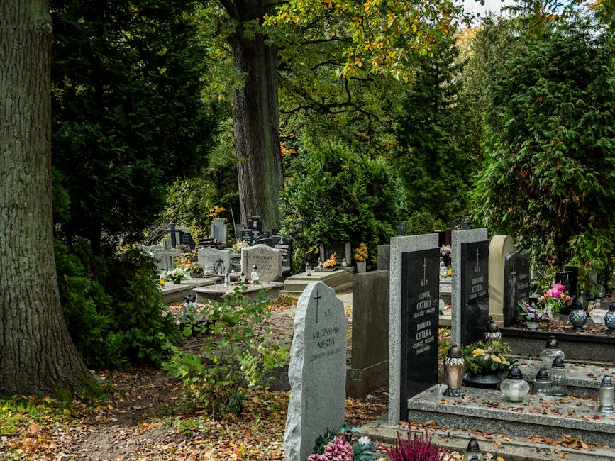 A view of a peaceful cemetery with several gravestones made of polished grey and black granite, some engraved with names and dates. The graves are decorated with floral arrangements, including pink, y