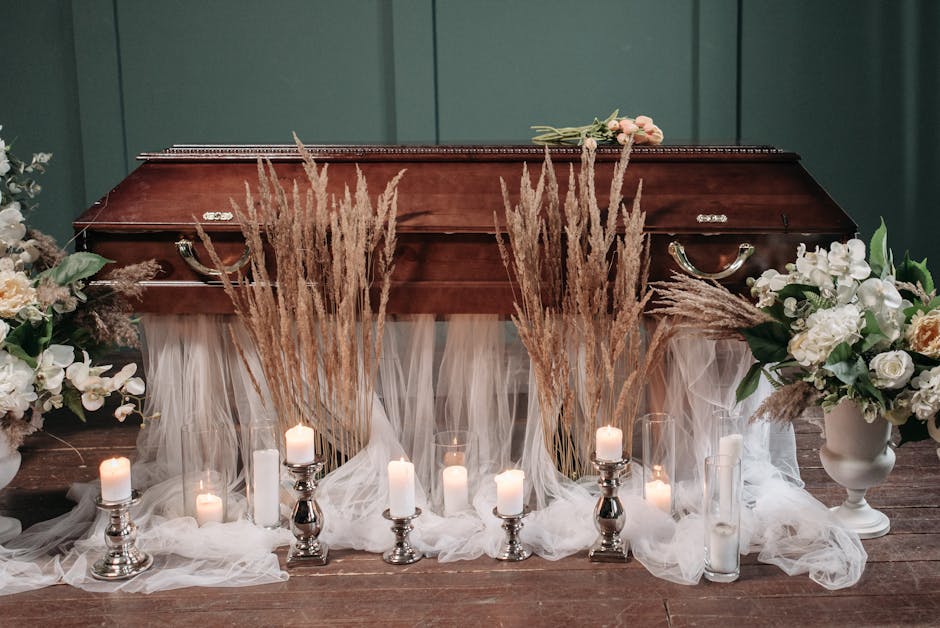 A funeral floral memorial arrangement placed in front of a polished wooden casket, featuring tall, beige dried pampas grass and white roses, lilies, and other white blooms in elegant white ceramic vas