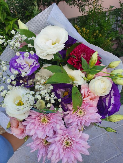 A vibrant bouquet of fresh flowers, including large pink dahlias, white lisianthus, purple and white eustoma, and red chrysanthemums, accented with delicate baby's breath and lush green foliage. The a