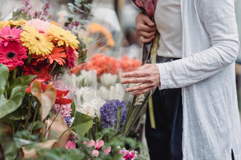 A person in a white cardigan and dark pants is selecting fresh flowers at a florist's stall, with a variety of vibrant blooms including pink, yellow, orange, white, purple, and red flowers arranged in