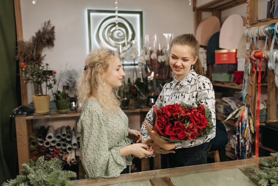 A florist with curly blonde hair wearing a light green patterned dress hands a vibrant bouquet of red roses, small red berries, and green foliage to a young woman with straight brown hair styled in a 